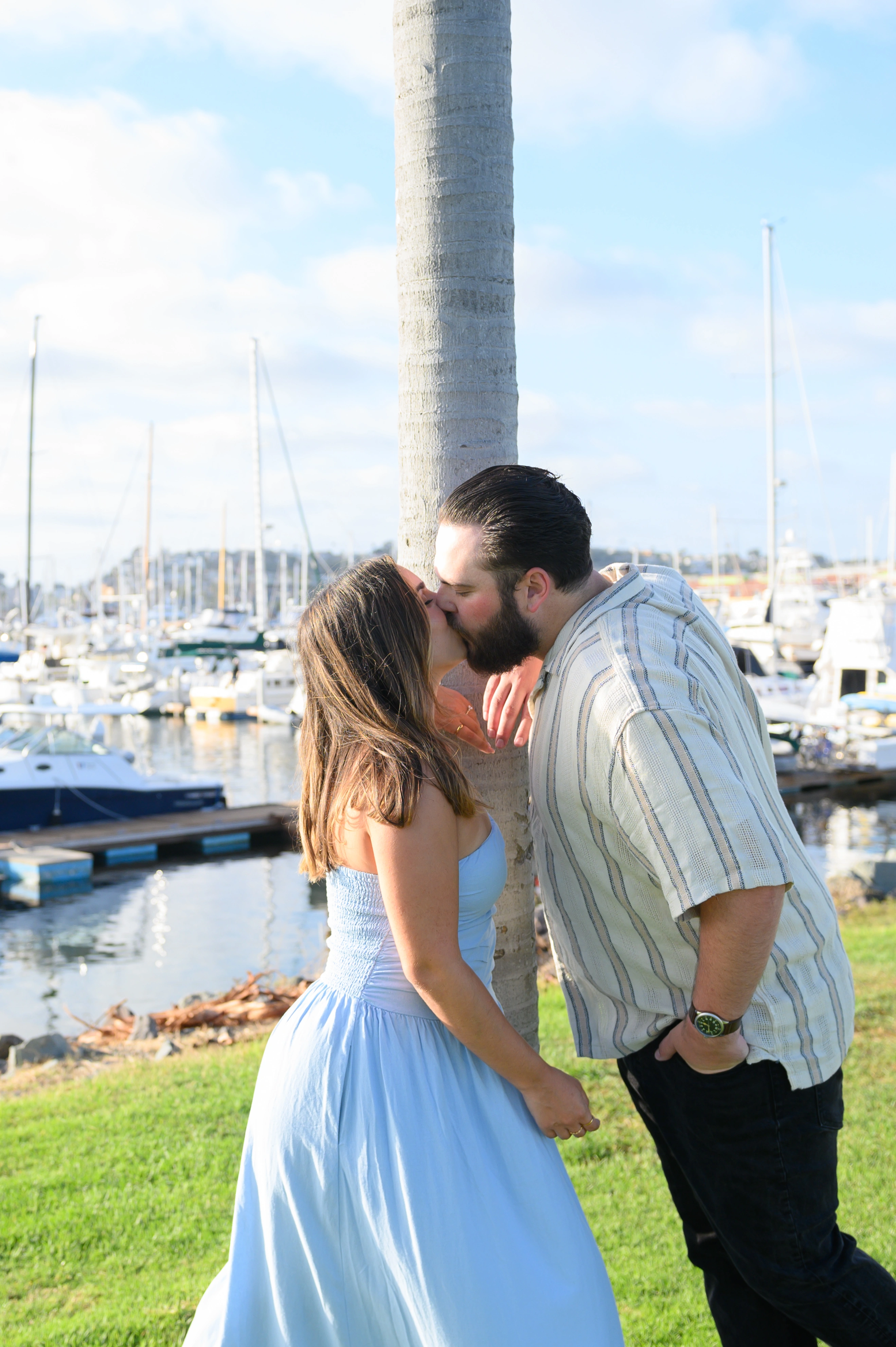 Romantic couple sharing a kiss near a palm tree at a San Diego marina, captured by a San Diego proposal photographer.
