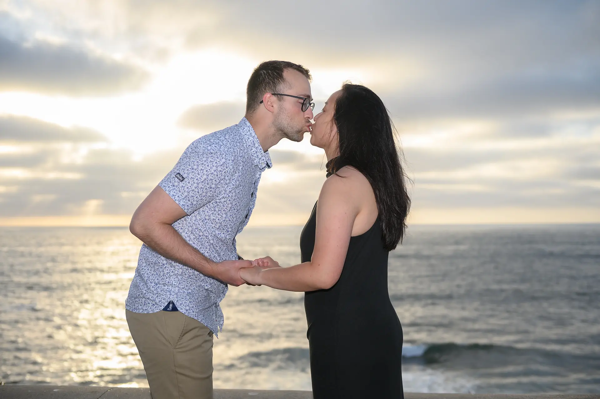The couple shares a tender moment, holding hands and leaning in for a kiss as the sun sets over the ocean.