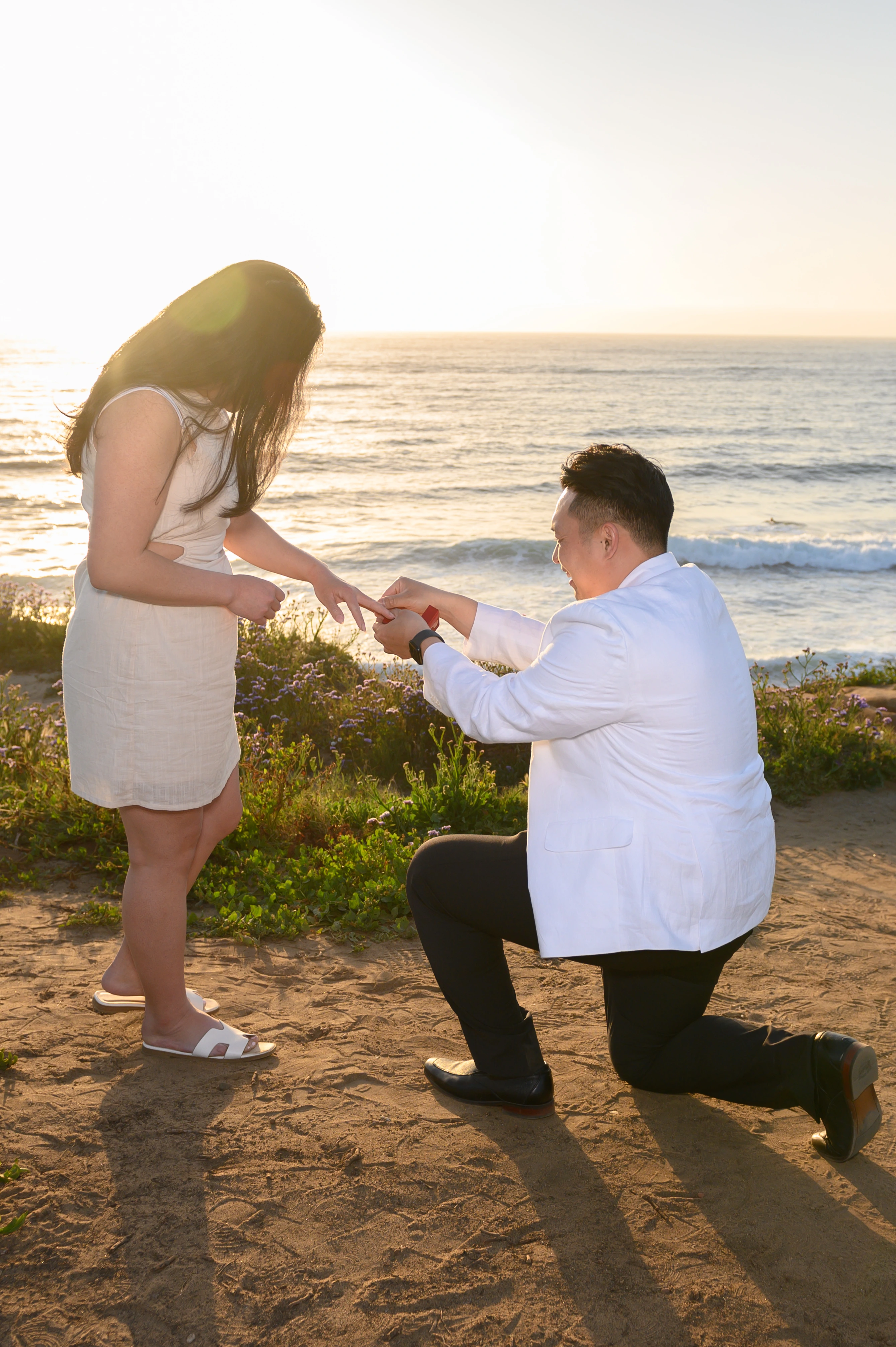A man kneels on one knee to propose to a woman on a beach at sunset, with the ocean in the background.