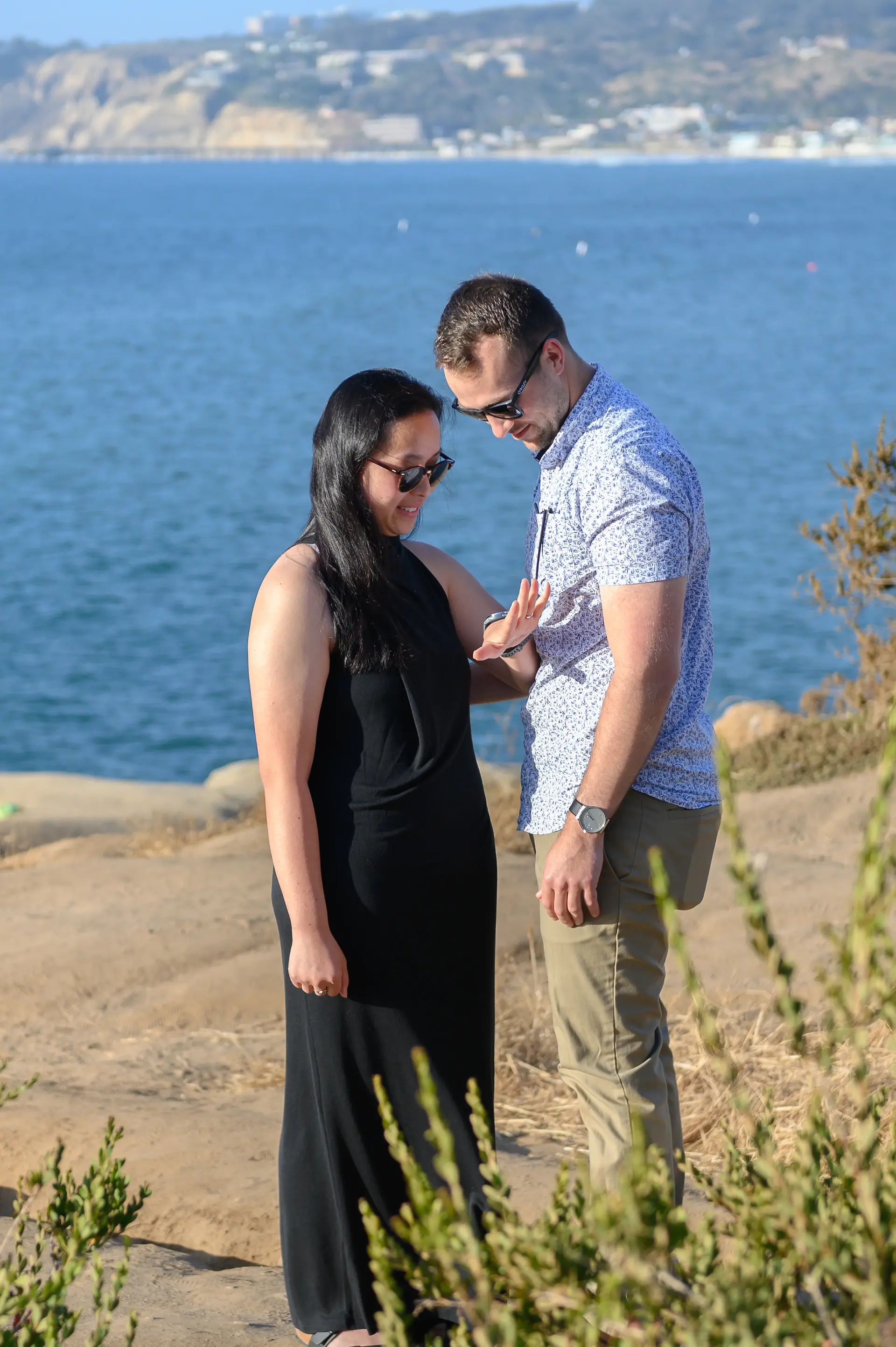 The couple stands on a rocky surface, with the woman showing her engagement ring against a scenic coastal backdrop