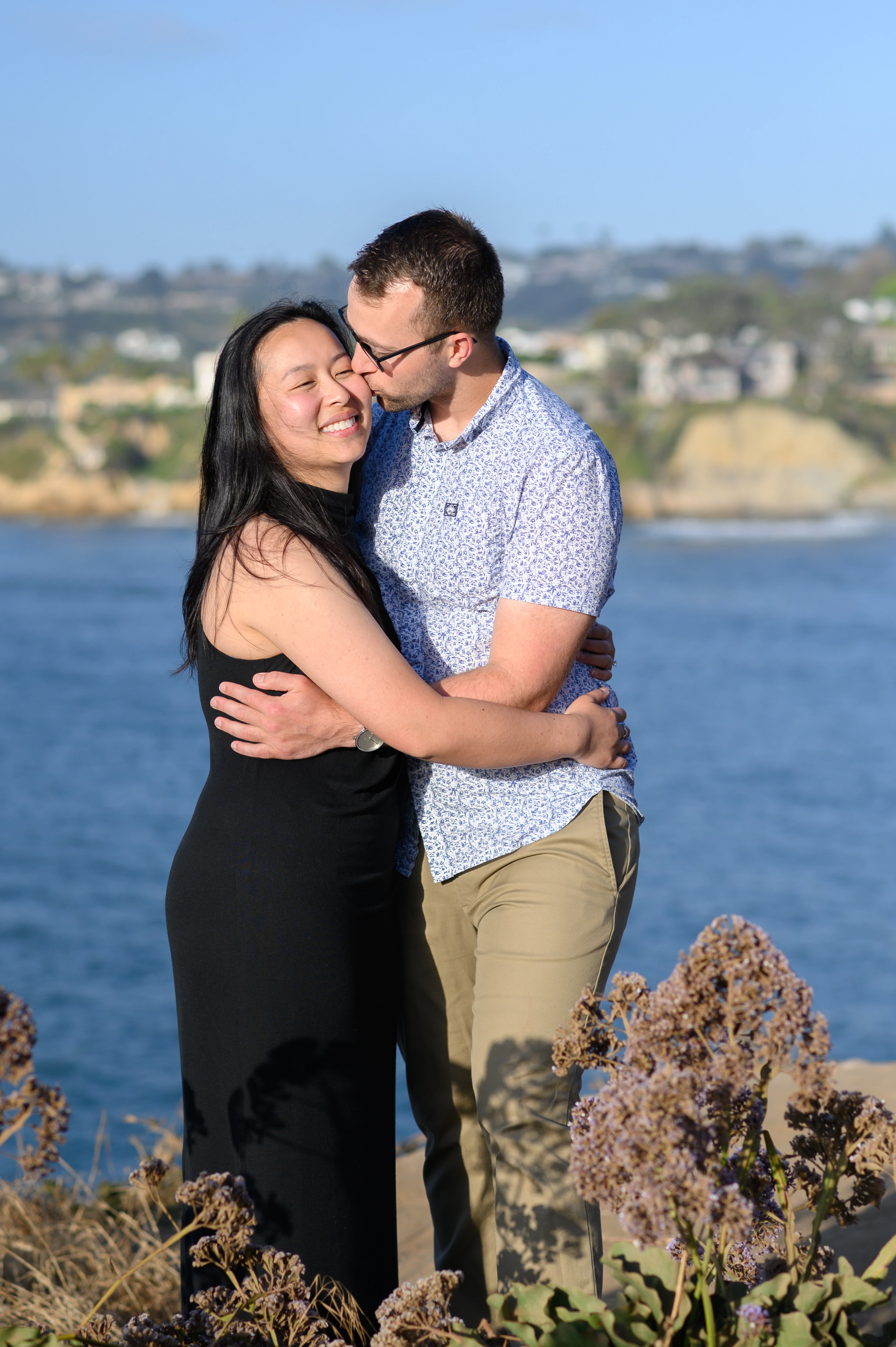 A couple embraces on a rocky shoreline with the ocean in the background, enjoying a moment together.