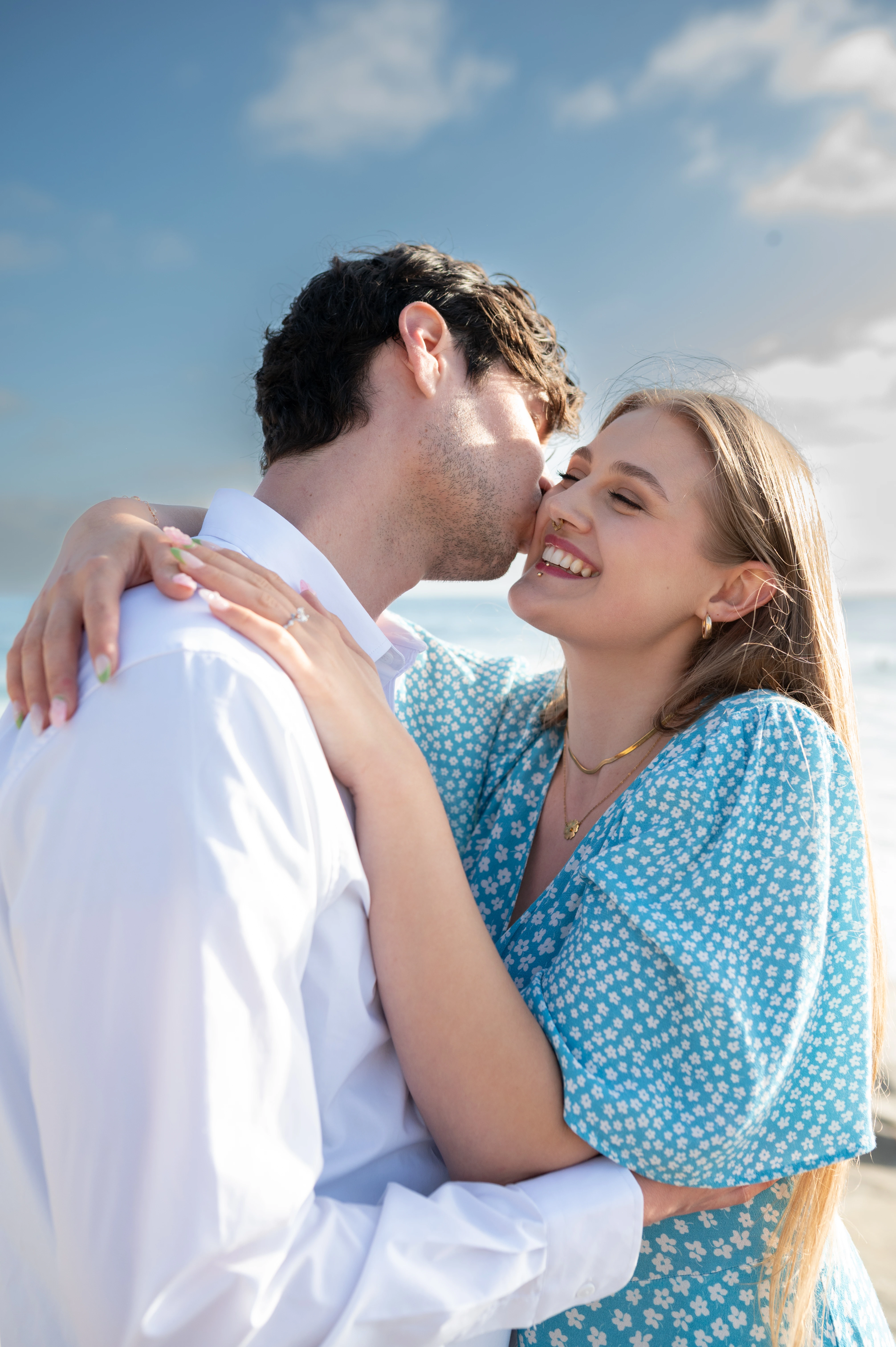 The couple shares a romantic kiss, framed by a bright blue sky and ocean waves