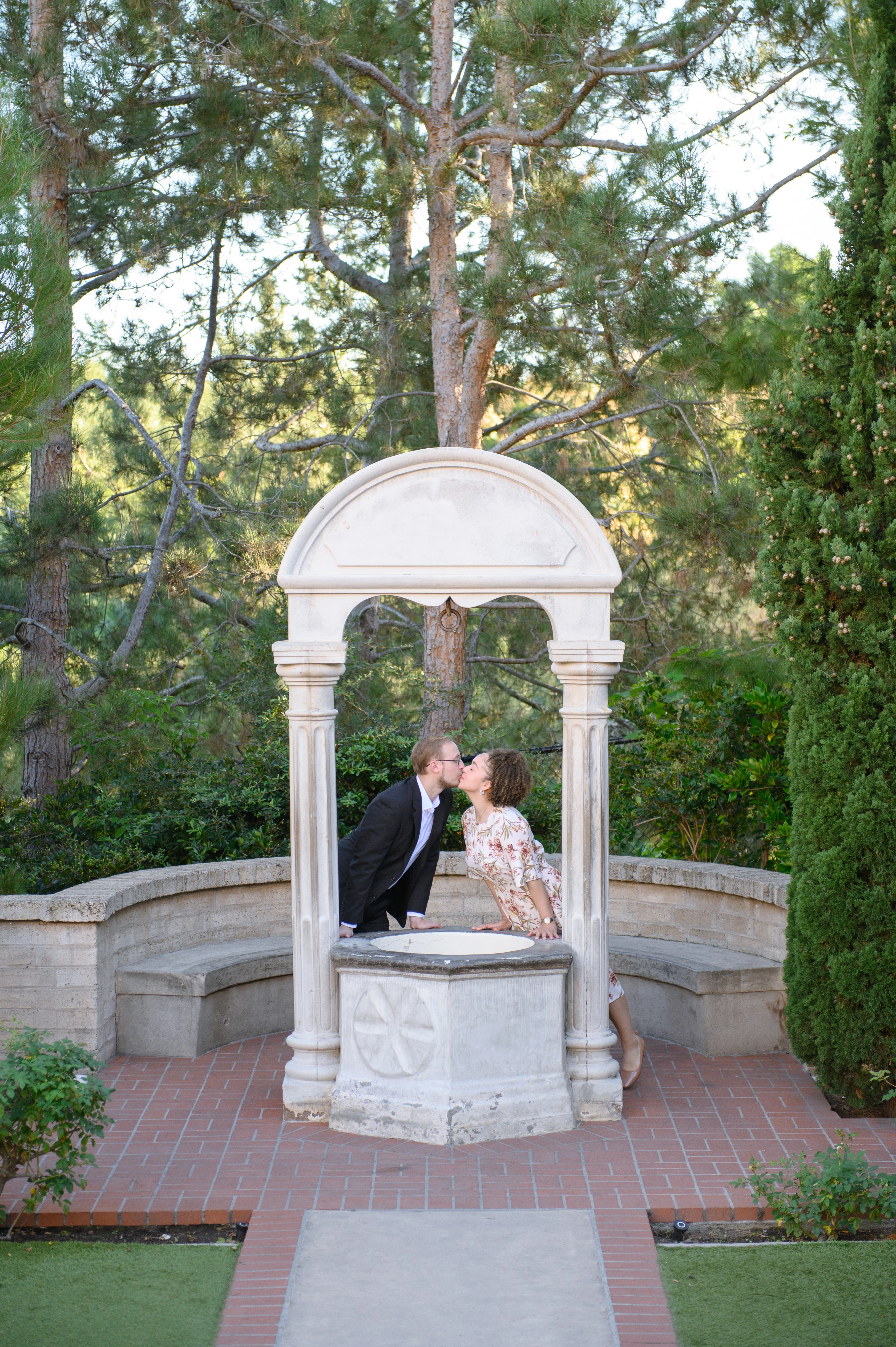 The couple shares a kiss under a charming stone gazebo, framed by lush greenery and tall trees
