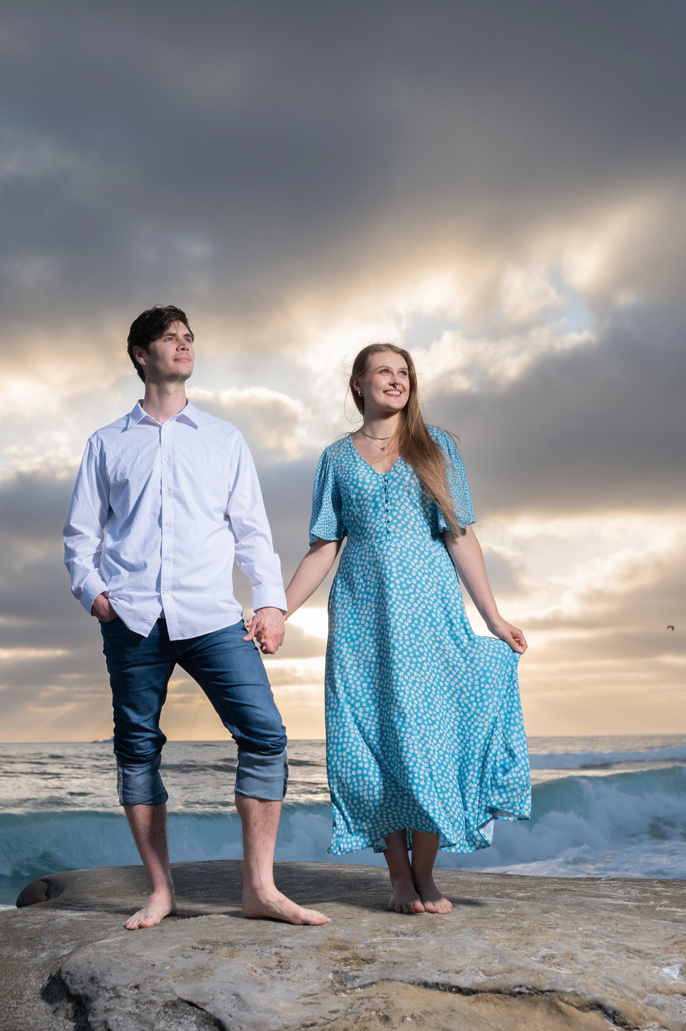 A couple stands hand in hand on a rocky beach, with a dramatic sky and ocean waves in the background