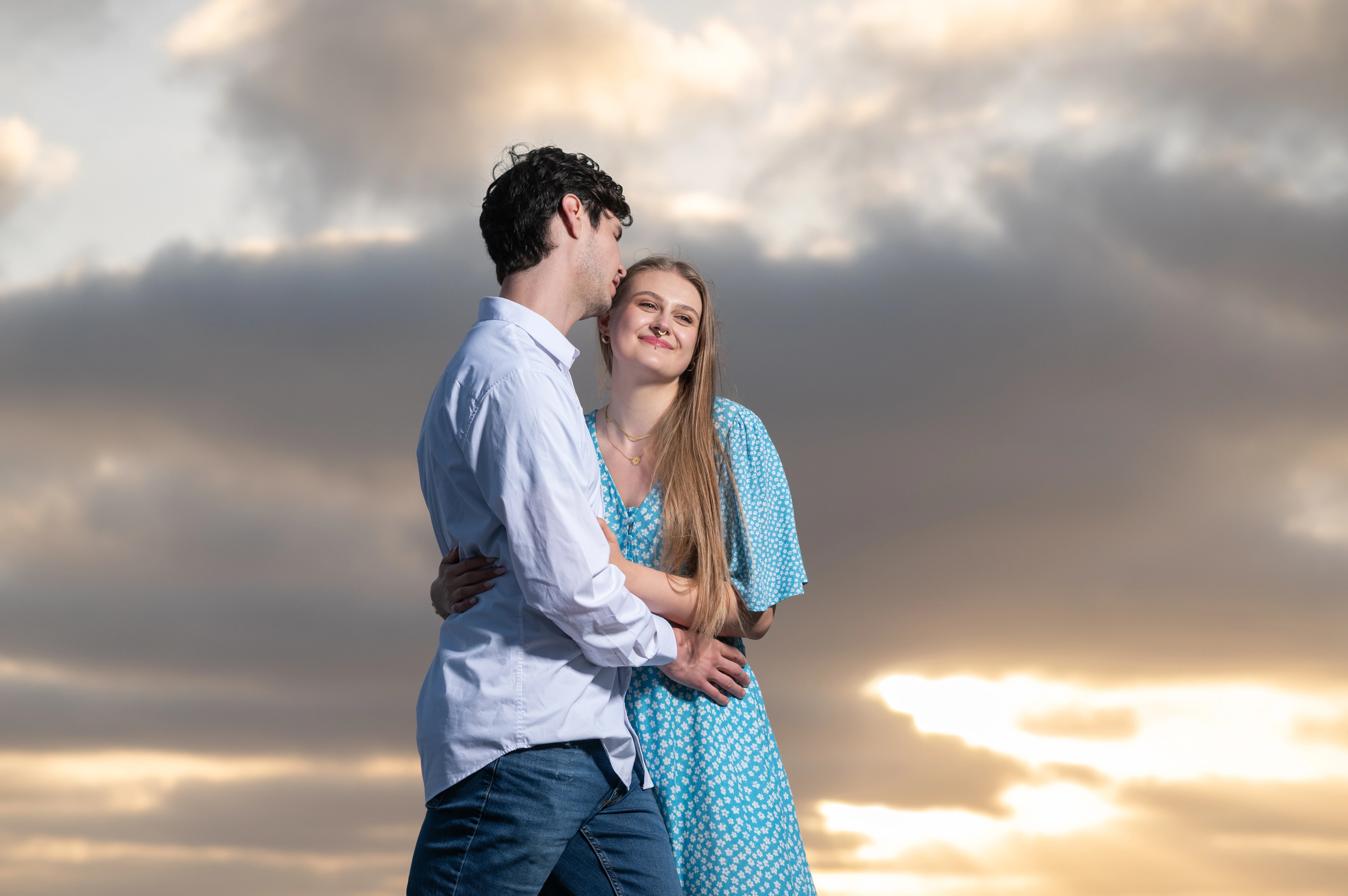 The couple embraces against a backdrop of a dramatic sunset, showcasing their affection in a serene setting