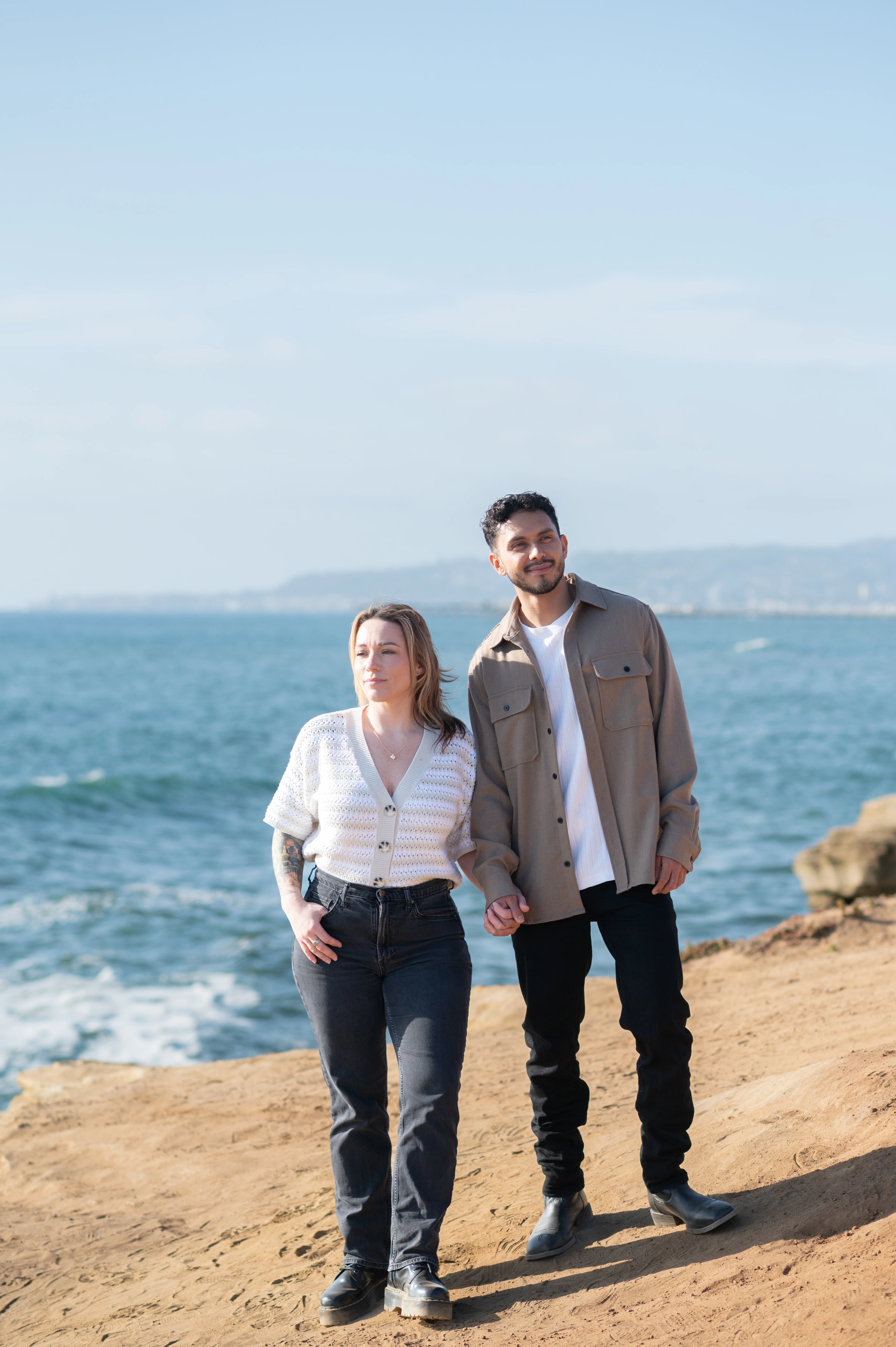 A joyful couple walks hand-in-hand along the sandy cliffs of Sunset Cliffs, with the sparkling ocean in the background. Their laughter and connection reflect the excitement of their engagement