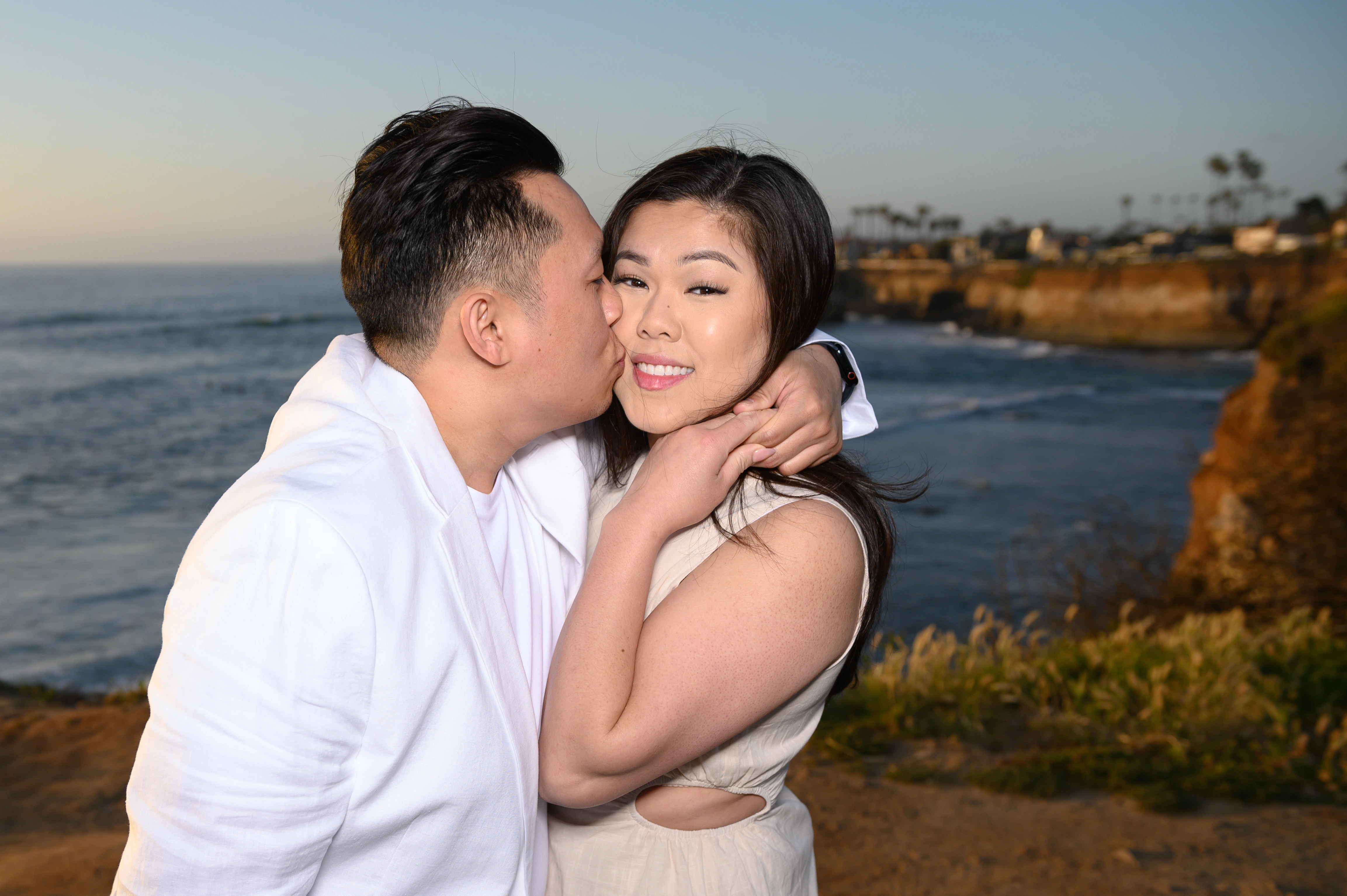 A couple shares a romantic moment by the ocean at sunset, with cliffs and palm trees in the background