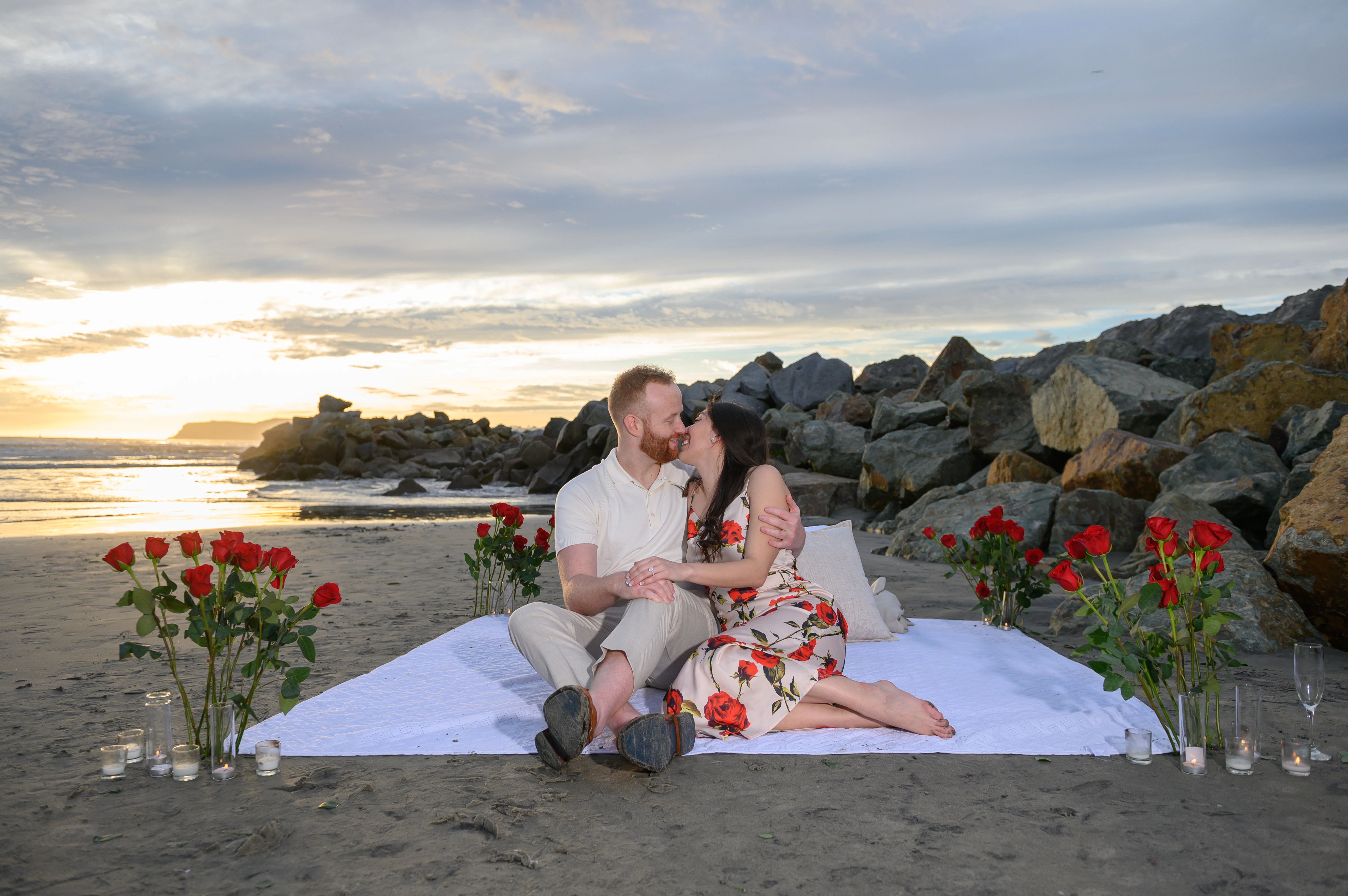 The couple shares a romantic kiss while seated on a blanket, framed by a backdrop of rocks and a beautiful sunset.