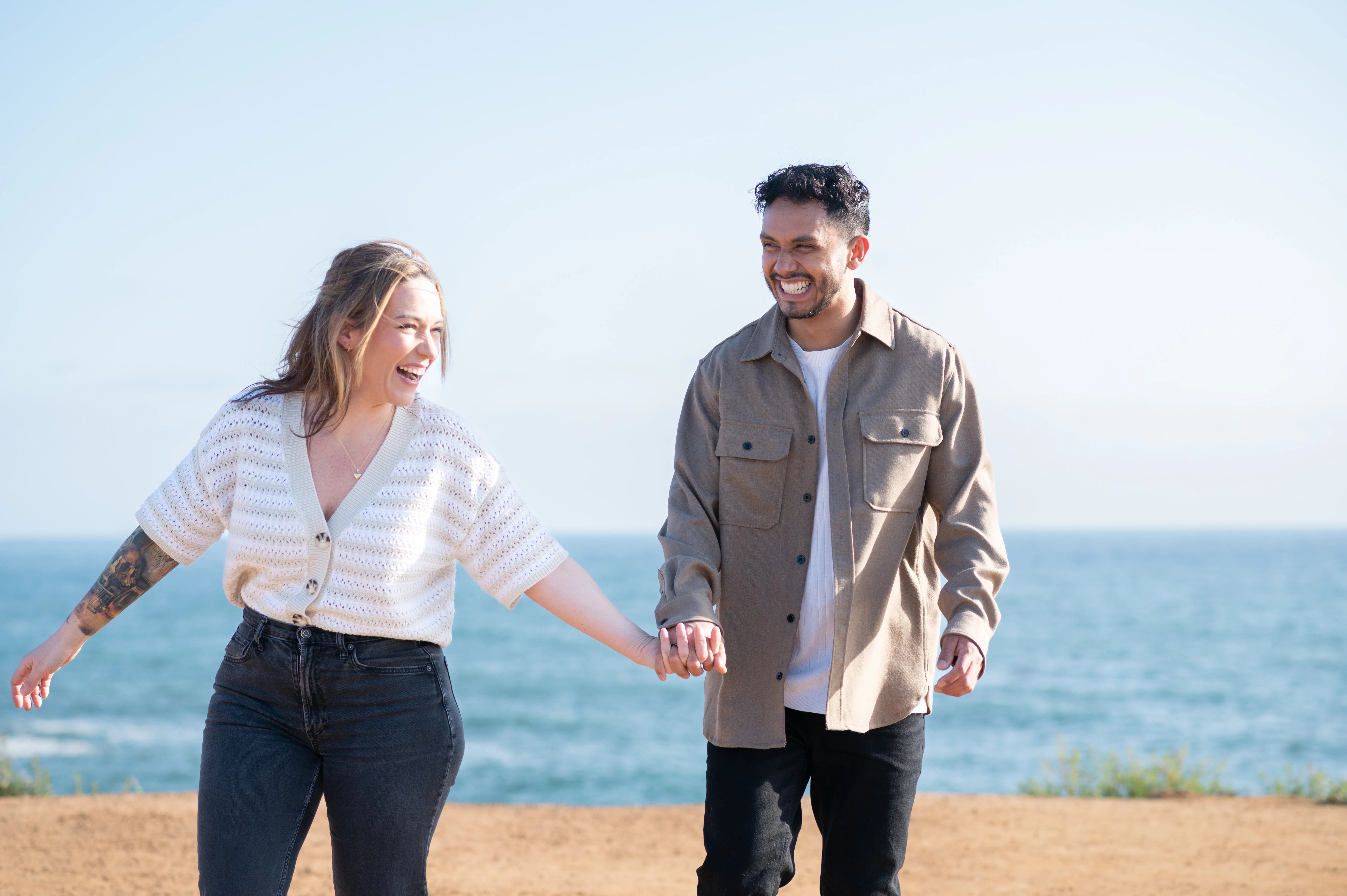 The couple walks hand-in-hand along the scenic coastline of Sunset Cliffs, laughing and radiating happiness. The clear sky and sparkling ocean enhance the romantic atmosphere of their engagement shoot