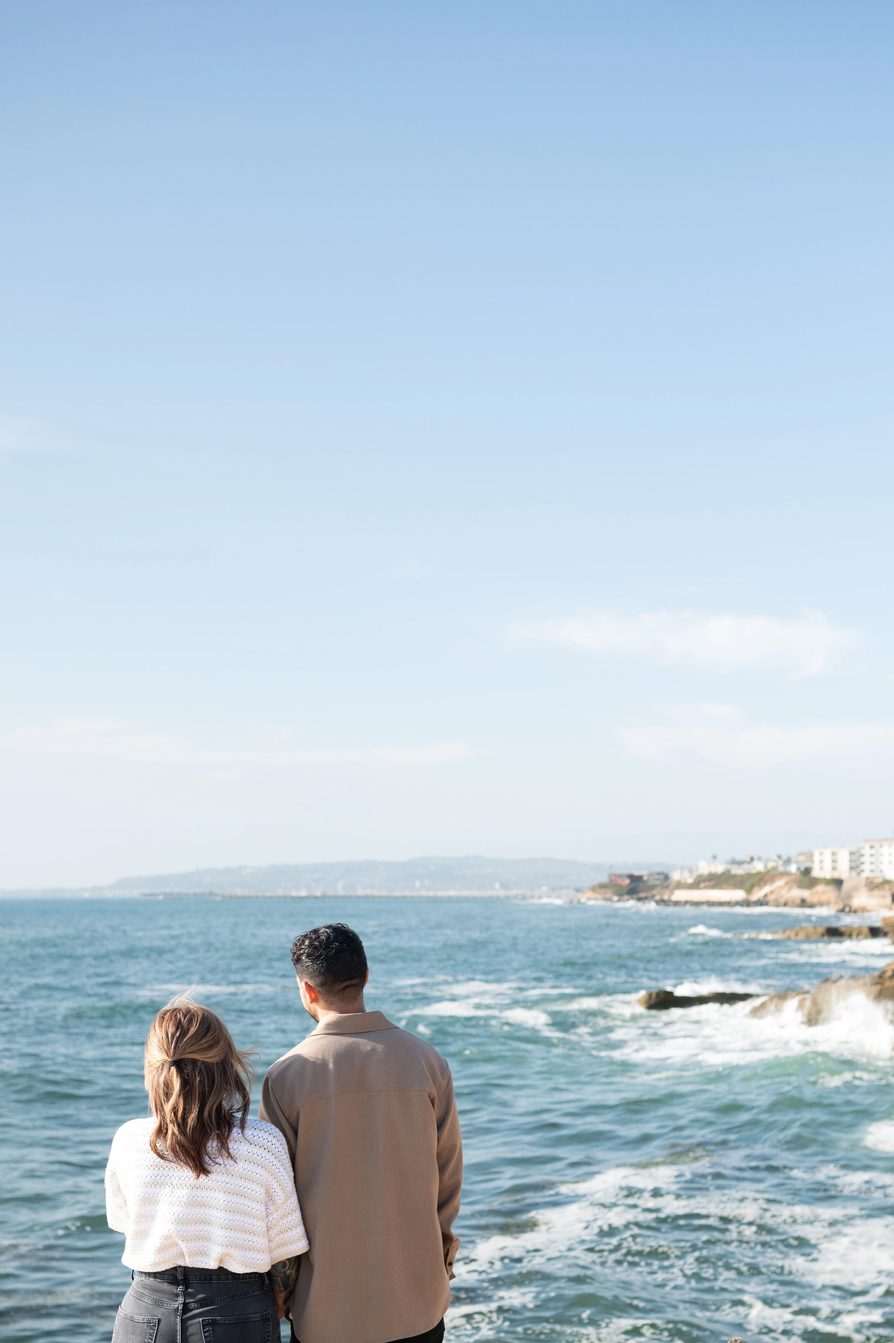 A couple stands hand in hand at Sunset Cliffs, gazing out at the serene ocean waves under a clear blue sky, capturing a moment of connection and anticipation during their engagement session.