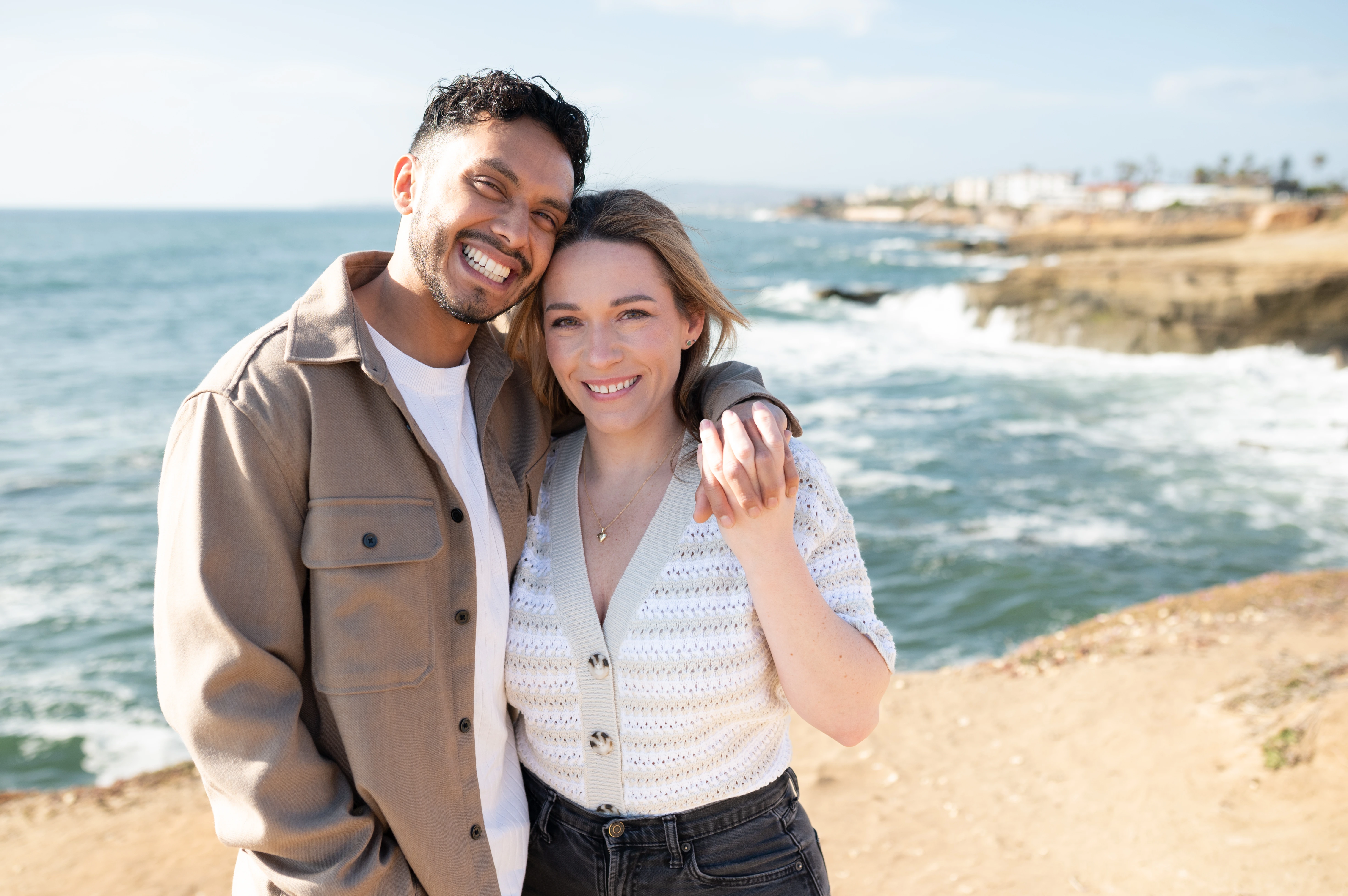 A close-up of a couple embracing at Sunset Cliffs, with the ocean waves gently crashing behind them. The woman shows off her engagement ring, symbolizing their love and future together