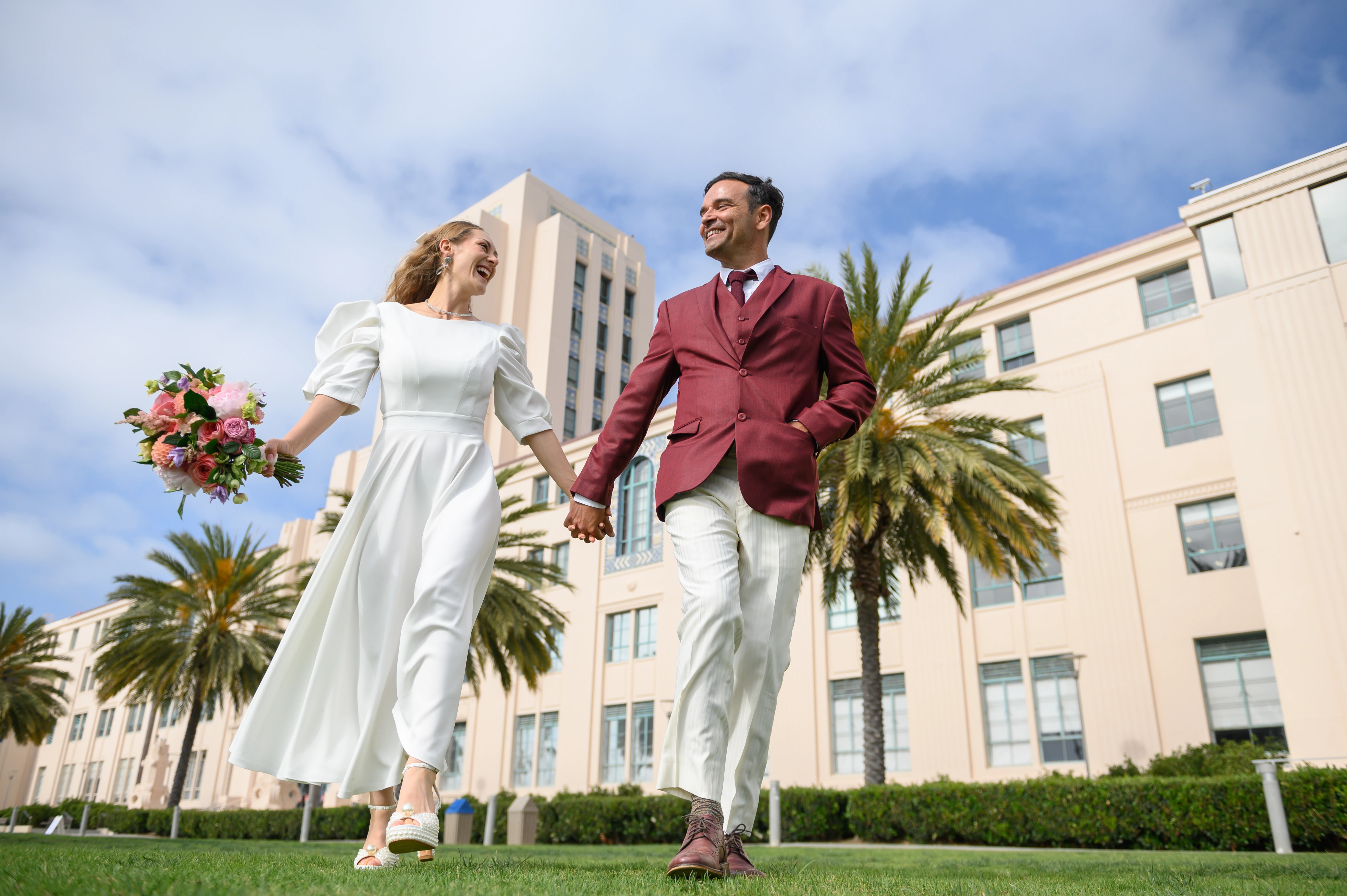 A couple holding hands and running together in front of a courthouse in a park, smiling and looking at each other affectionately