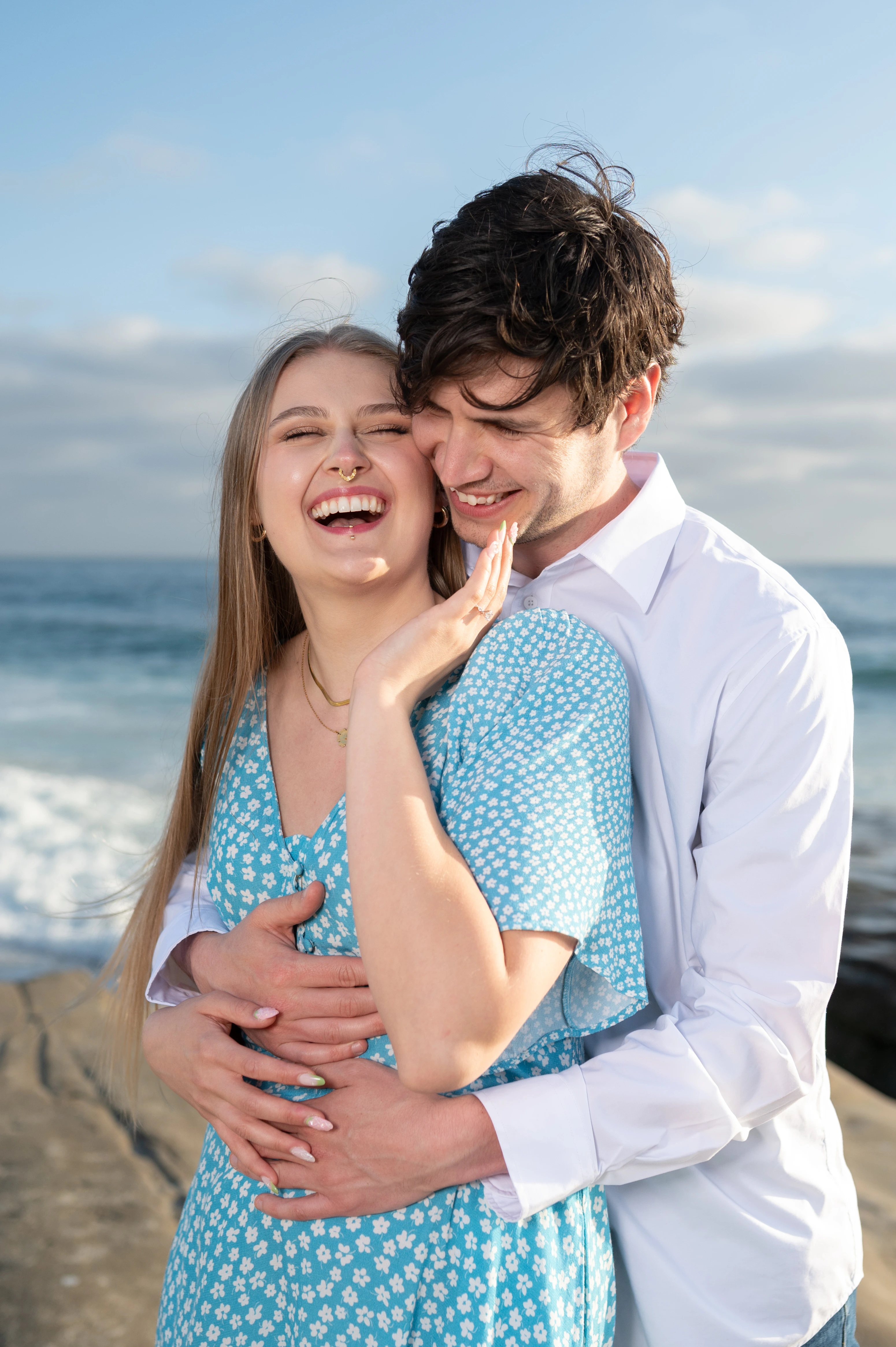 The couple embraces warmly, with the ocean visible in the background, capturing a moment of intimacy