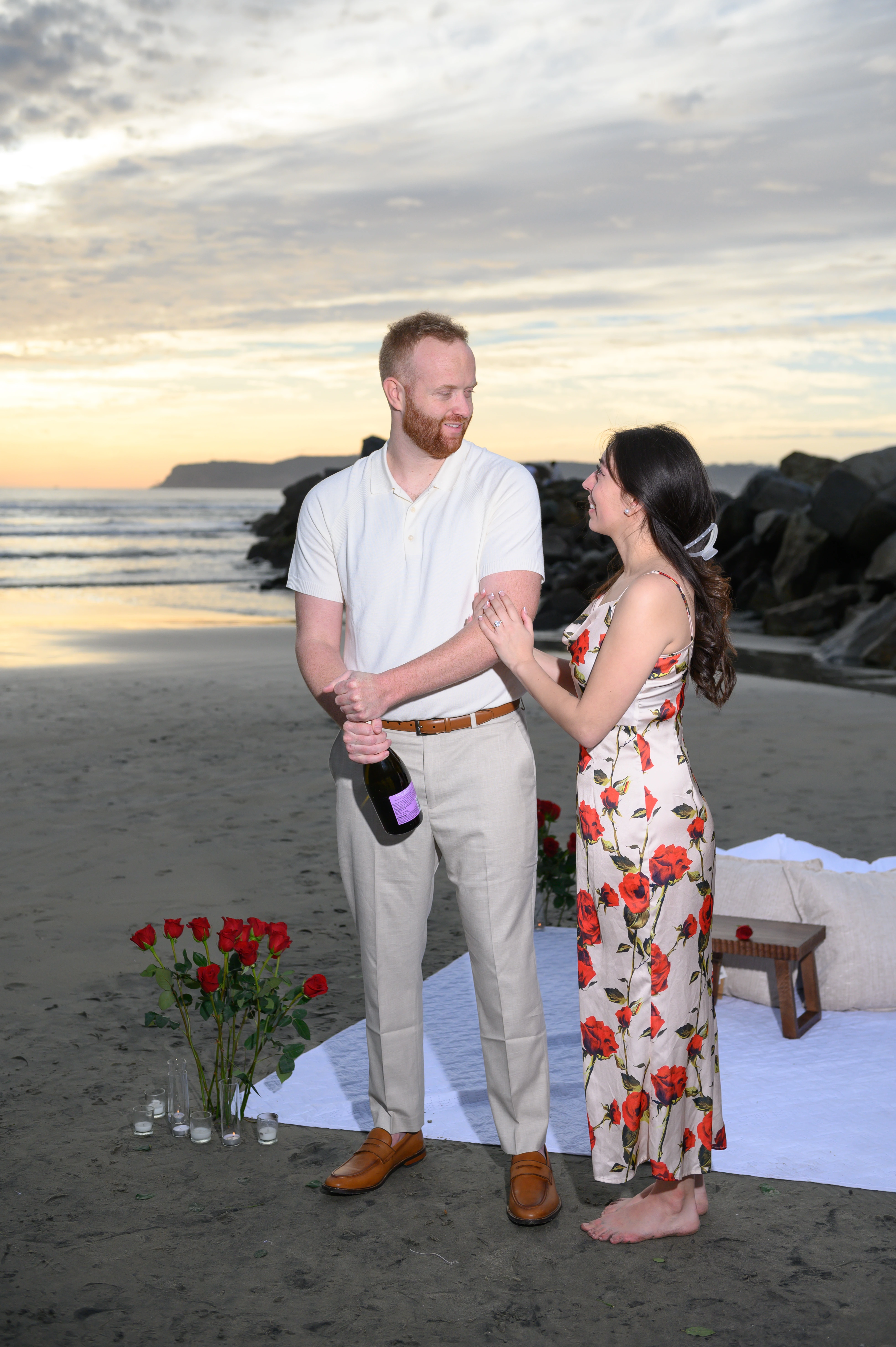 A couple stands on the beach at sunset, surrounded by roses and a picnic setup, with one person holding a champagne bottle