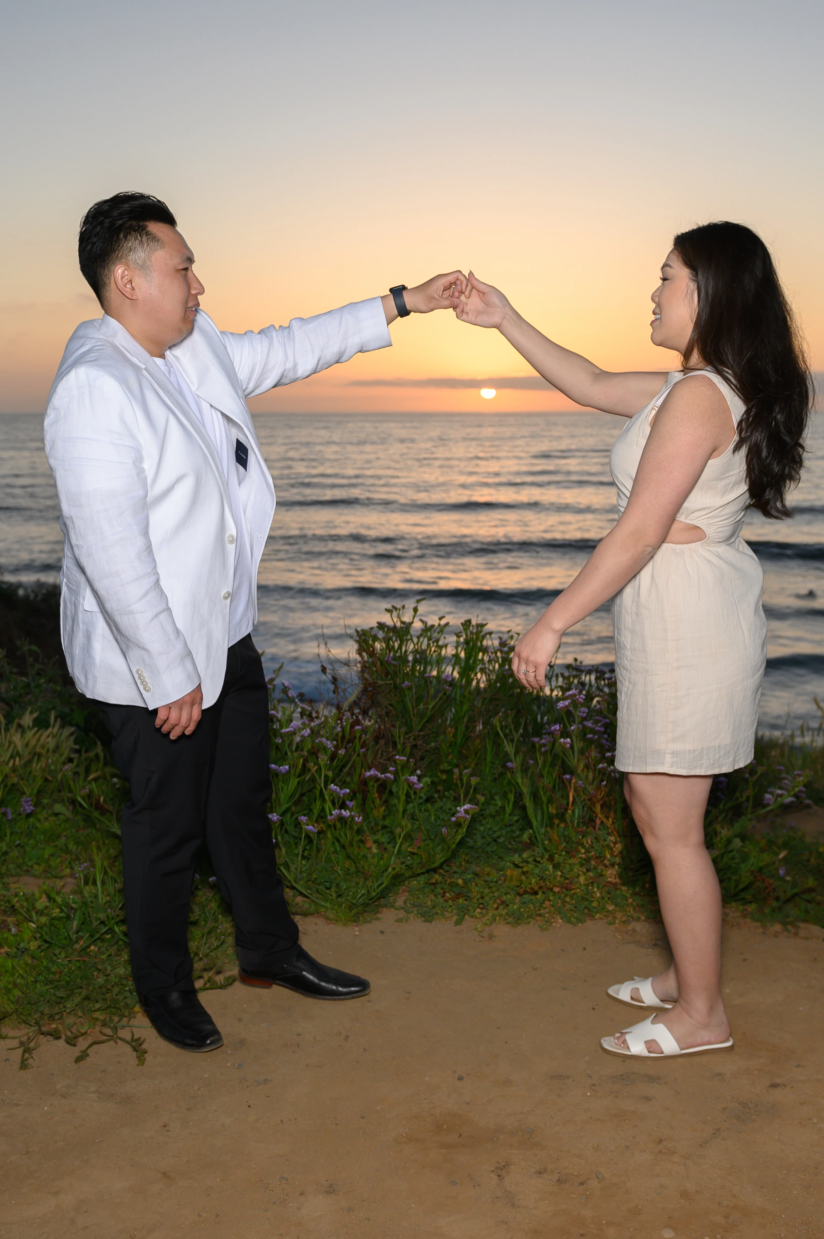 A couple stands on a beach at sunset, holding hands as the sun sets over the ocean. The scene captures a romantic moment with vibrant colors in the sky and gentle waves in the background.