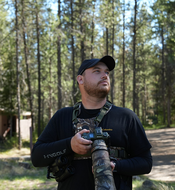 A photo of a tall birder with a beard, dark hat, and dark long sleeve shirt. He's holding a large camera covered in camera