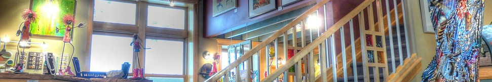 Interior view of Osmosis Gallery featuring whimsical mixed-media sculptures, vibrant paintings, and a central staircase with wooden railings. Natural light streams through large windows, highlighting the artwork on display.