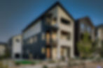 Contemporary three-story residence at Baseline with a steep gable, mixed light and dark panels, second-floor balcony, third-floor covered deck, and warm interior light at dusk.