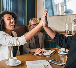 Women laughing and giving a hi-five across a dining table.