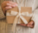 Hands holding a brown gift box with a white lace ribbon. Background features a beige knitted sweater, creating a warm, cozy mood.