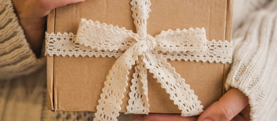 Hands holding a brown gift box with a white lace ribbon. Background features a beige knitted sweater, creating a warm, cozy mood.