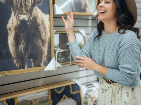 A woman wearing a hat laughs while looking at cow artwork in a store. She wears a blue sweater and floral skirt. Art pieces feature cows.