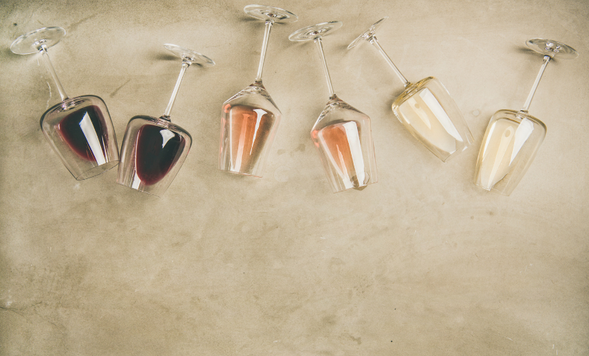 Five wine glasses tipped over on a beige surface, filled with red, rosé, and white wine in a gradient, creating a casual, artistic feel.