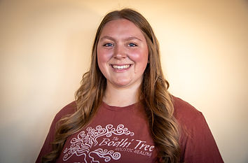 A picture of a blonde woman smiling. She is wearing a maroon shirt advertising The Bodhi Tree, a holistic spa.