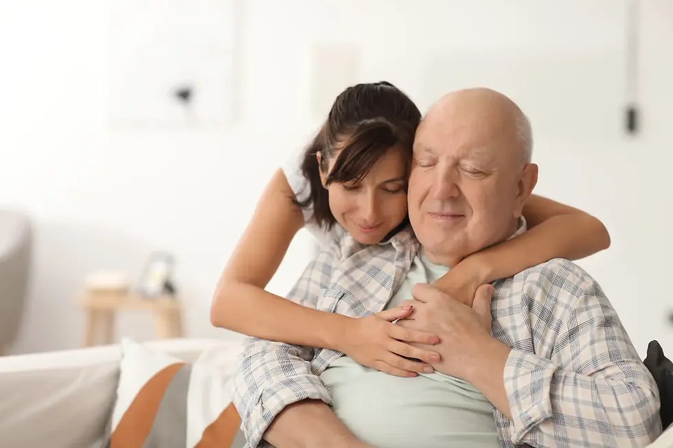 Adult daughter hugging her elderly father from behind as he sits on a sofa with eyes closed, both smiling peacefully.