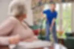 A friendly female carer in a blue uniform vacuuming a bright, well-kept living room while smiling at an elderly woman, representing coordinated home care services as part of a brokered care package.