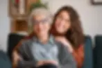 Smiling elderly woman sitting comfortably on a sofa with her adult daughter embracing her from behind, representing positive outcomes from personalised care planning and family-supported decision-making.