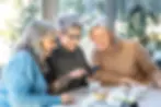Three smiling women enjoying coffee and cake while looking at a smartphone together, symbolising positive outcomes and improved quality of life following a comprehensive care review process.