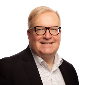 Hymyilevä silmälasipäinen vaaleatukkainen mies profiilikuvassa. Katse kameraan, puku päällä, valkoinen kauluspaita.