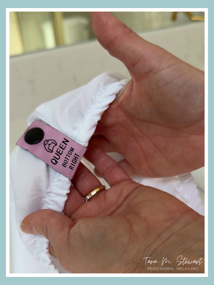 A close up of hands holding a white fitted sheet with a pink snap-on label that reads Queen Bottom Right; a simple solution for identifying sheet sizes in a linen closet.