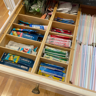 Open drawer with neatly organized compartments containing drink mixes and straws. Wooden dividers and labels for items.