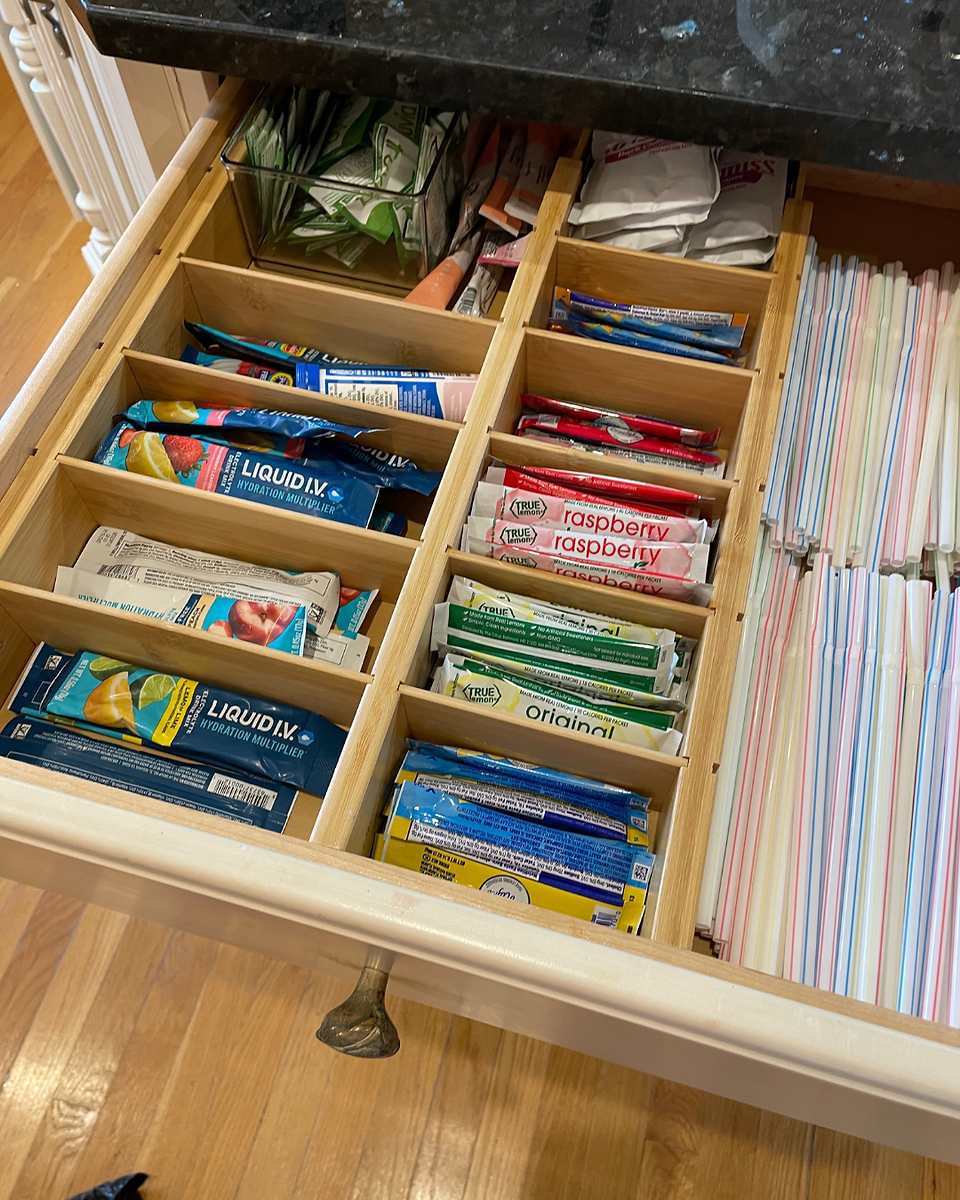 Open drawer with neatly organized compartments containing drink mixes and straws. Wooden dividers and labels for items.