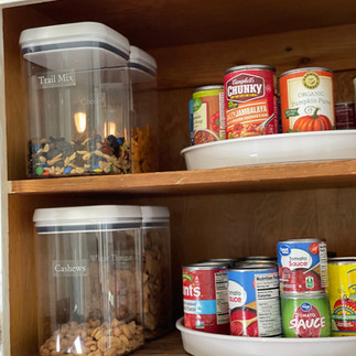 Kitchen cabinet organization featuring airtight containers for trail mix and cashews with subtle white-on-clear labels and a turntable for canned goods.