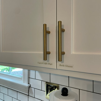 White kitchen cabinets featuring subtle white-on-clear labels for "Cups & Coffee Mugs," "Rocks Glasses,"