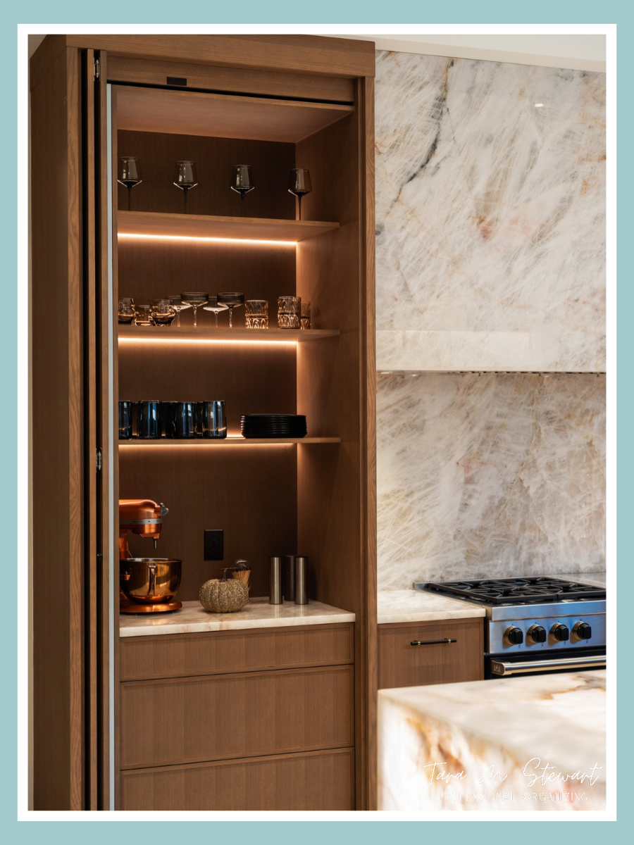 Custom oak appliance garage cabinet designed by Jimmy Nash Homes with sliding pocket doors, integrated LED shelf lighting, stacked glassware, black dishware, and a copper stand mixer set up by Tara M. Stewart Professional Organizing.