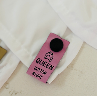 A close-up view showing neat hands holding a white bed sheet with a clean, tan snap-on label that clearly displays "Queen" and "Bottom Right" for easy size identification in a linen closet