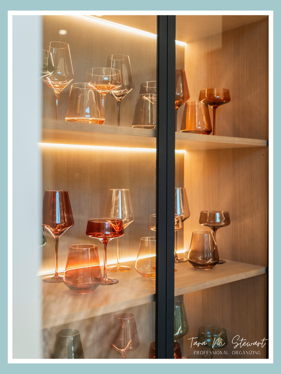 Custom built-in glass cabinetry with warm LED shelf lighting featuring amber wine stems and colorful glassware styled by height and tone by Tara M. Stewart Professional Organizing.