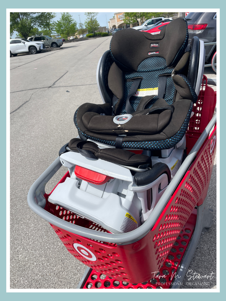 A red plastic shopping cart in a large store parking lot. The main part of the cart is holding a grey car seat base, and a black and teal car seat is balanced carefully on top, ready for drop-off.