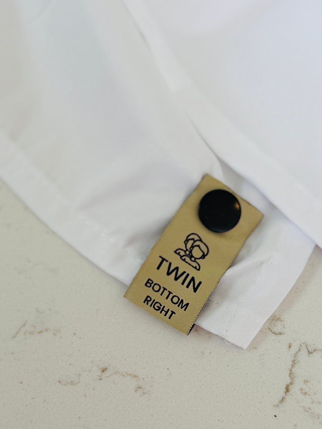 A close-up view showing neat hands holding a white bed sheet with a clean, tan snap-on label that clearly displays "Twin" and "Bottom Right" for easy size identification in a linen closet