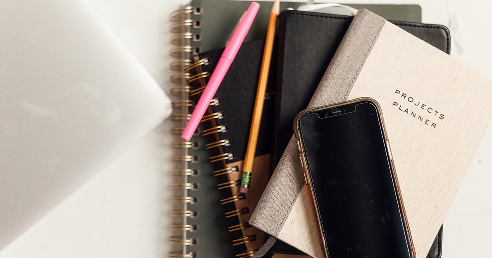 A flat lay view of a clean, organized white workspace featuring a neat stack of planners and notebooks, a pink pen, a yellow pencil, a modern smartphone, and the edge of a sleek laptop.