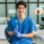 Smiling dentist in blue scrubs holds a dental mirror, seated in a clinic with dental equipment. Bright, clean setting.