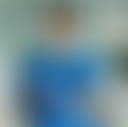 Smiling dentist in blue scrubs holds a dental mirror, seated in a clinic with dental equipment. Bright, clean setting.