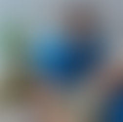 Medical professional in blue scrubs aids a soldier on a couch, administering an injection. Army backpack nearby, shelves and plants in the background.