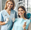 Dentist in blue scrubs smiles holding dental tools next to a patient in a dental chair with a blue bib, both looking cheerful.