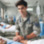 A man in an Air Force uniform with a stethoscope smiles, holding a patient's hand in a hospital room with medical equipment.