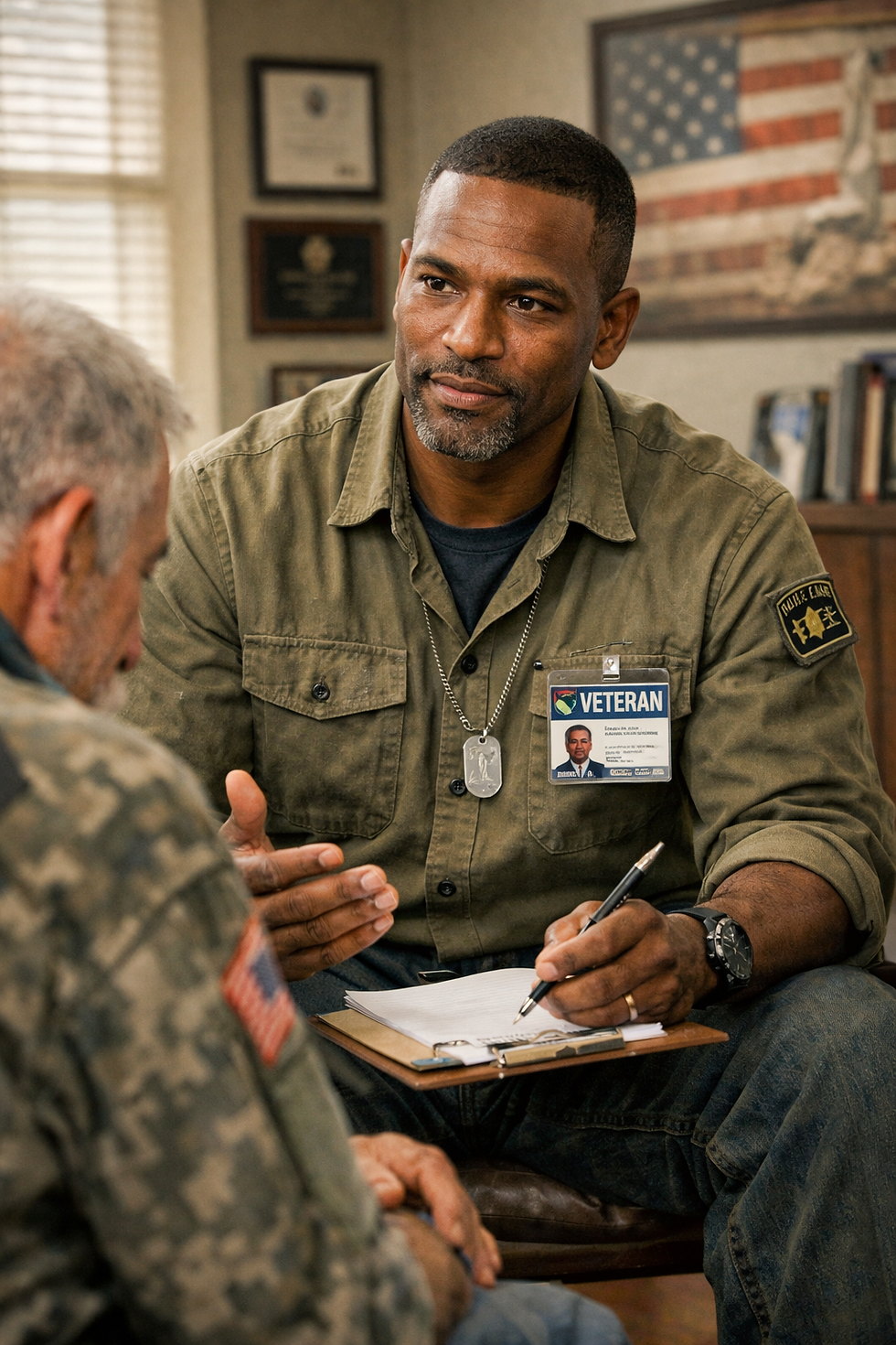 Black veteran in olive shirt writes on a clipboard while speaking with an older man in a cozy office with an American flag.